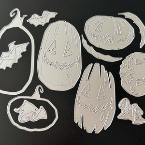 May include: A collection of silver-colored metal cutting dies featuring various Halloween-themed shapes, including three distinct jack-o'-lantern pumpkin faces, two bat silhouettes, and two pumpkin outlines, all designed for paper crafting projects.