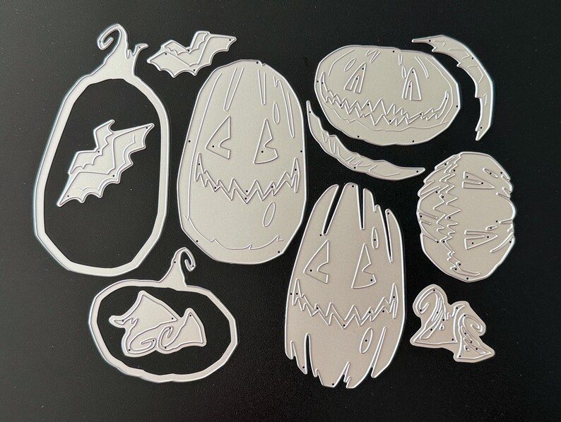 May include: A collection of silver-colored metal cutting dies featuring various Halloween-themed shapes, including three distinct jack-o'-lantern pumpkin faces, two bat silhouettes, and two pumpkin outlines, all designed for paper crafting projects.