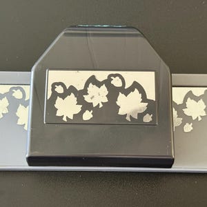 May include: A black and gray craft paper punch featuring a repeating cutout pattern of maple leaves and small acorns. The tool has a wide rectangular base with a central raised handle section that displays the leaf and acorn design in silver.