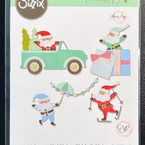 May include: A Sizzix Thinlits die set, model 665958 by Olivia Rose, featuring 11 metal cutting dies. The packaging displays die-cut shapes including a light green truck with a Santa figure, two gift boxes, a Santa skiing, and a Santa with an umbrella.