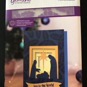 May include: A Gemini embossing folder set featuring a Christmas card design. The card has a blue background with a nativity scene and the text "Joy to the World." The card measures 5" x 7" (12.7cm x 17.8cm).