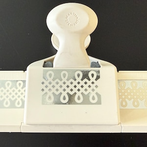 May include: A cream-colored paper craft punch with a decorative silver design. The punch features a handle with the text "MARTHA STEWART" and a design that creates a repeating pattern. Ideal for scrapbooking and paper crafts.