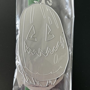 May include: A silver-colored metal craft die in the shape of a tall, carved pumpkin with a jagged, toothy grin and triangular eyes. The die is enclosed in a clear plastic storage bag with a green zipper seal at the top for protection.