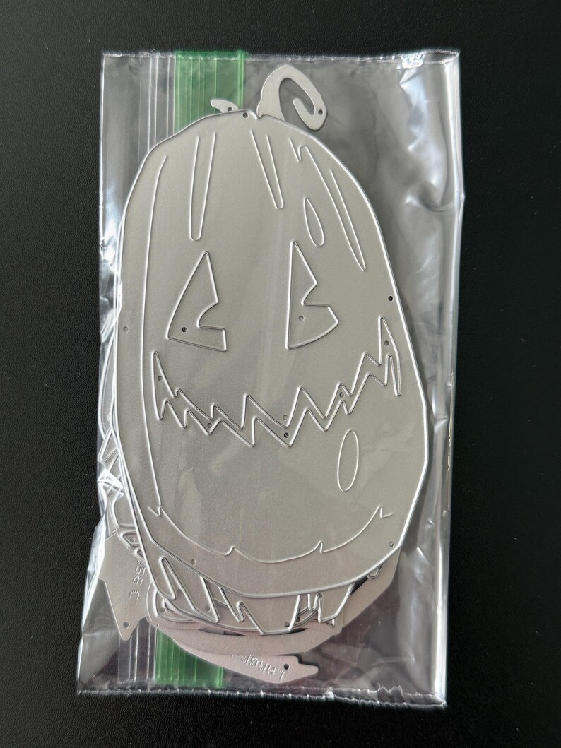 May include: A silver-colored metal craft die in the shape of a tall, carved pumpkin with a jagged, toothy grin and triangular eyes. The die is enclosed in a clear plastic storage bag with a green zipper seal at the top for protection.