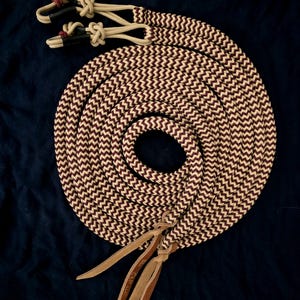 May include: A coiled rope with a chevron pattern in cream and burgundy. The rope has leather accents and knotted ends. The background is a dark blue fabric. The rope is likely used for equestrian activities.