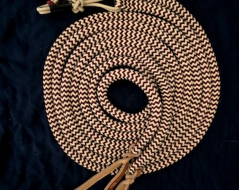 Peut inclure: Une corde enroulée avec un motif en chevrons crème et bordeaux. Elle présente des accents en cuir et des extrémités nouées. Le fond est un tissu bleu foncé. La corde est probablement utilisée pour l'équitation.