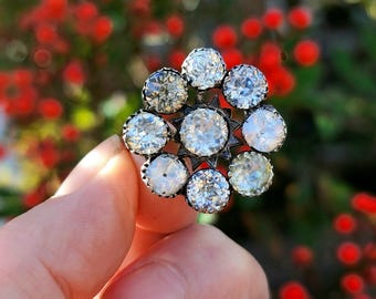 May include: A silver-toned ring featuring a floral design with nine clear, sparkling gemstones. The central stone is larger, surrounded by eight smaller stones. The ring is held against a blurred background of red berries and green foliage.