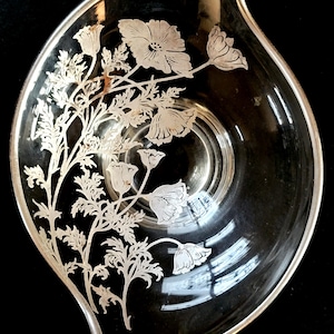 Op de afbeelding: Transparante glazen serveerschaal met een zilveren rand en bloemenmotief. De schaal heeft een asymmetrische vorm en is voorzien van geëtste bloemen en bladeren. Het ontwerp bevindt zich aan de linkerkant van de schaal. De schaal staat op een zwarte achtergrond.