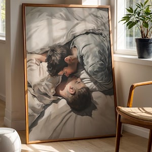 May include: A framed artwork depicts two people in a tender embrace, lying under a white blanket. The artwork is displayed in a well-lit room with a wooden chair, a potted plant, and a small white stool. The room has a neutral color palette.