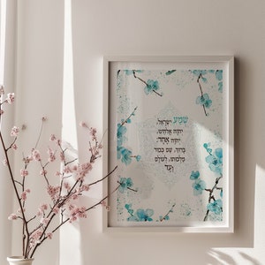 May include: A white framed print with a watercolor design of blue flowers and a Hebrew text that reads "Shema Yisrael, Adonai Eloheinu, Adonai Echad. Baruch Shem Kevod Malchuto, Le'olam Va'ed." The print is hanging on a white wall with a vase of pink flowers and a stone on a white shelf below.