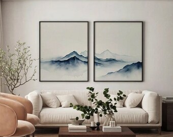 Puede incluir: Dos pinturas de acuarela enmarcadas de montañas azules con niebla blanca en un salón. Las pinturas están colgadas en una pared blanca sobre un sofá blanco y una mesa de café con un jarrón de plantas verdes.