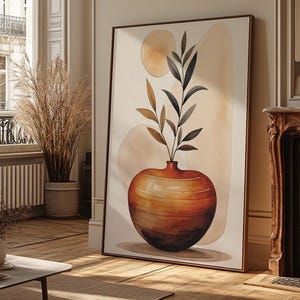 May include: A framed abstract art print featuring a large, orange vase with dark leaves. The artwork has a neutral color palette with beige and brown tones. The print is leaning against a wall in a room with hardwood floors and a window.