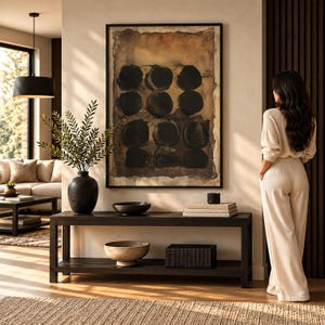 May include: A rectangular black wooden console table with two tiers, featuring a black vase with green foliage, a dark bowl, a stack of books, and a black box. Above the table hangs a large framed abstract painting with black circles.