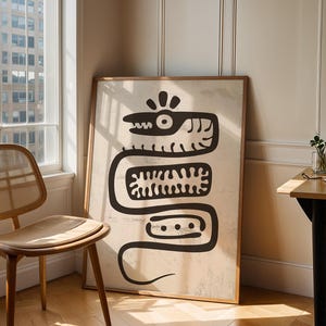 May include: A framed art print featuring a stylized black snake design on a beige background. The artwork is displayed in a room with a wooden chair, a small table, and a window with natural light.
