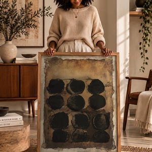 May include: A rectangular framed wall art piece featuring a textured, earthy brown background with a grid of nine solid black circles. The artwork is encased in a light-toned wooden frame with a visible grain, showcasing a distressed, aged paper aesthetic.