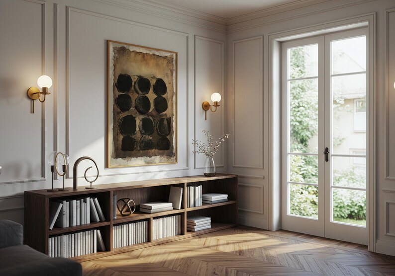 May include: A low, rectangular wooden bookshelf with multiple compartments filled with white books, topped with decorative metal sculptures and a glass vase, positioned below a framed abstract painting with black circles on a brown background.