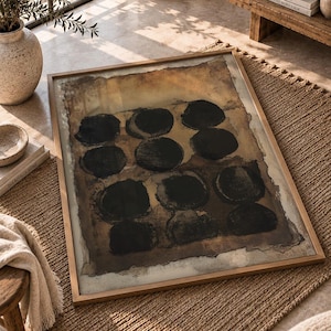 May include: A rectangular framed wall art piece featuring a series of twelve dark black circular shapes arranged in a grid pattern against a textured, mottled brown and tan background with uneven, deckled edges within a thin light wood frame.