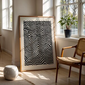 Puede incluir: Una impresión artística enmarcada con un patrón ondulado en blanco y negro. La obra de arte se exhibe en una habitación luminosa con una silla de madera, una planta en maceta y un pequeño taburete blanco. El marco es de color marrón cálido.