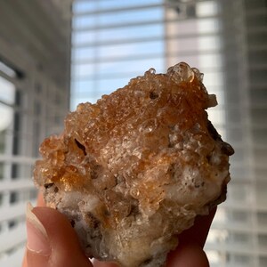 May include: A close-up of a cluster of orange and white crystals. The crystals have a bubbly, textured appearance and are set against a blurred background. The crystals are held in a hand.