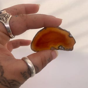 May include: A polished agate stone slice with a vibrant orange and yellow centre, framed by a darker, textured border. The stone is held between fingers, showcasing its natural patterns and colours. The agate is a unique, decorative item.