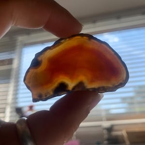 May include: A translucent, polished agate slice, held between fingers. The stone features bands of orange, red, and brown hues, with darker edges. The natural gemstone is illuminated by sunlight, showcasing its layered patterns and textures.