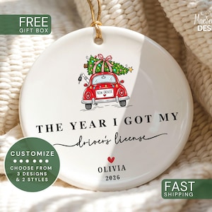 May include: A round white ceramic ornament featuring a red car illustration with a Christmas tree on top, a license plate reading NEW DRIVER, and the text THE YEAR I GOT MY driver's license, OLIVIA, and 2026 with a small red heart icon.