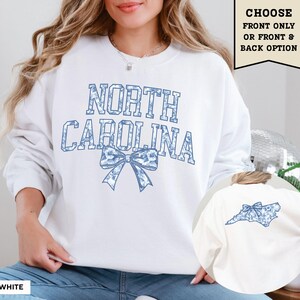 A woman wearing a white sweatshirt with the text "NORTH CAROLINA" printed on it, along with a blue bow design.
