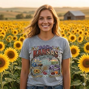 May include: A heather gray t-shirt featuring a colorful graphic print with the text She's From KANSAS I CAN TELL. The design includes a map of Kansas, a football helmet, a bison, a tornado, red shoes, wheat, a pumpkin, and sunflowers.