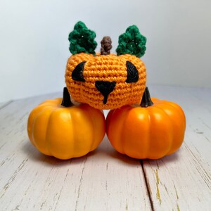 May include: A crocheted orange pumpkin with green leaves and black eyes sits on top of two orange plastic pumpkins.