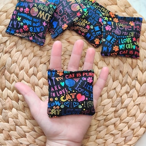 May include: A hand holding a small black cat toy with colorful text that says "My cat is purrfect, I love my cat, are in love, I little meow, cat love, I love my cat".