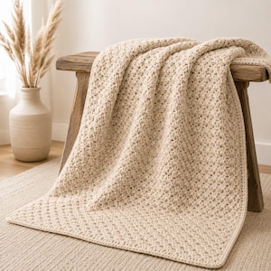Può includere: Una coperta all'uncinetto color beige con un motivo a punti ripetitivo e testurizzato. La coperta è drappeggiata su una panca in legno, mettendo in mostra la sua morbida struttura intrecciata e il colore marrone chiaro uniforme.