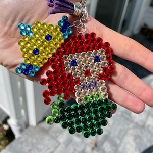 Puede incluir: Un llavero hecho a mano con una sirena pelirroja y un pez amarillo, ambos adornados con pedrer&iacute;a de colores. La sirena tiene una cola verde. El llavero incluye un anillo plateado y una borla morada.