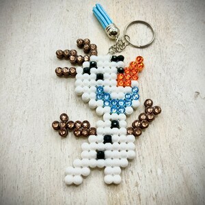 May include: A white, blue and brown beaded snowman keyring. The snowman is wearing a blue and white beaded scarf and has a brown beaded hat. The keyring is attached to a silver key ring.