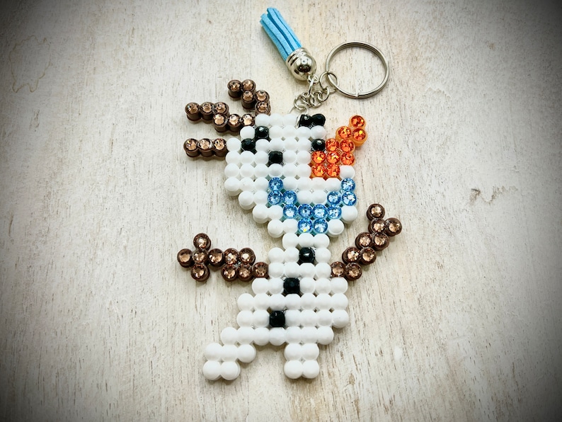 May include: A white, blue and brown beaded snowman keyring. The snowman is wearing a blue and white beaded scarf and has a brown beaded hat. The keyring is attached to a silver key ring.