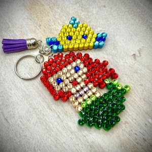Puede incluir: Un llavero hecho a mano con un cohete y una corona, ambos adornados con pedrer&iacute;a de colores. El cohete es rojo, blanco y verde, mientras que la corona es amarilla y azul. Una borla morada cuelga del llavero.