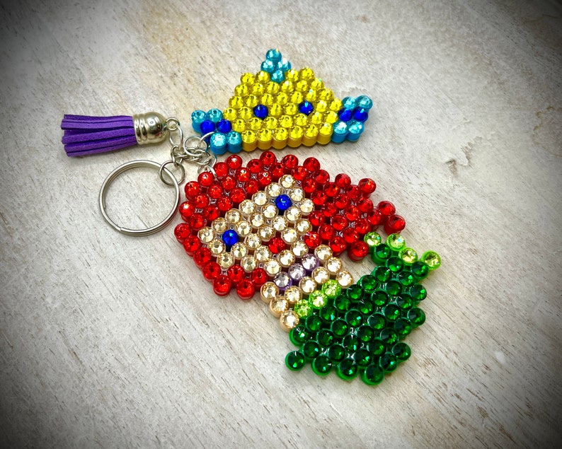 Puede incluir: Un llavero hecho a mano con un cohete y una corona, ambos adornados con pedrer&iacute;a de colores. El cohete es rojo, blanco y verde, mientras que la corona es amarilla y azul. Una borla morada cuelga del llavero.