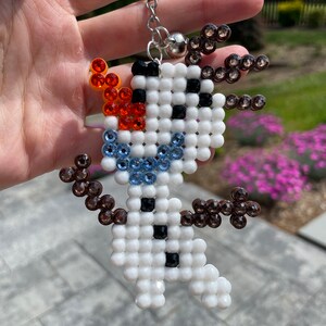 May include: A white beaded snowman keychain with brown, blue and orange beads. The snowman has a black button and a black smile.