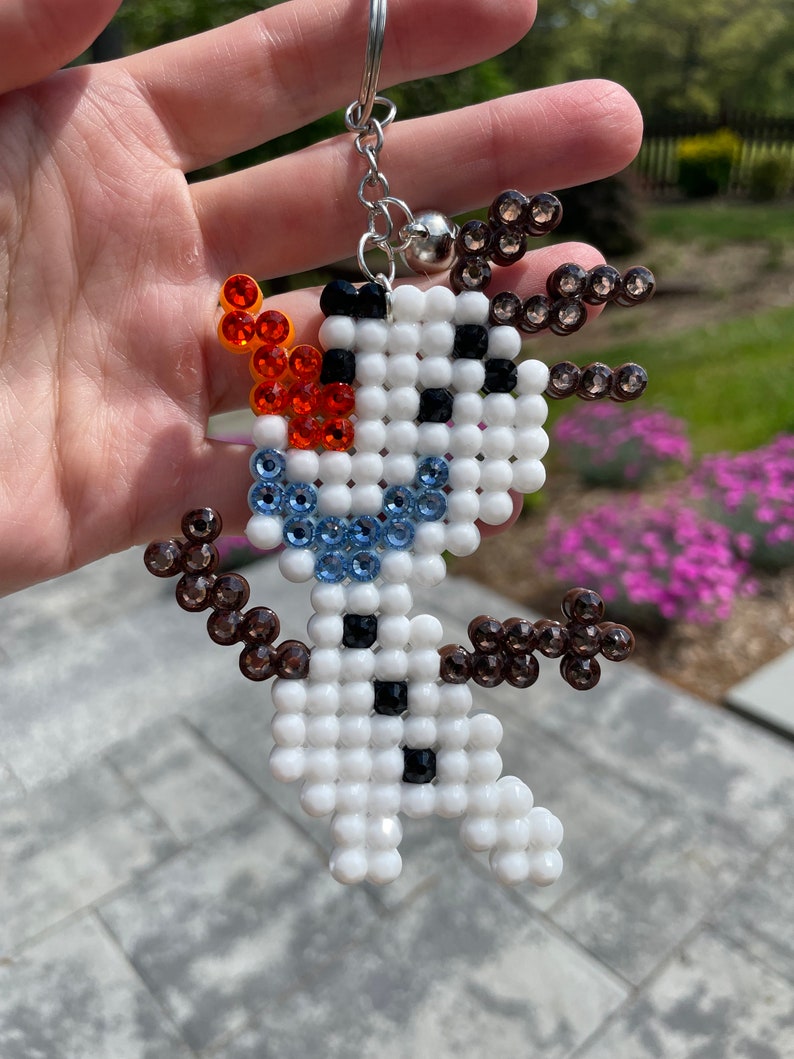 May include: A white beaded snowman keychain with brown, blue and orange beads. The snowman has a black button and a black smile.