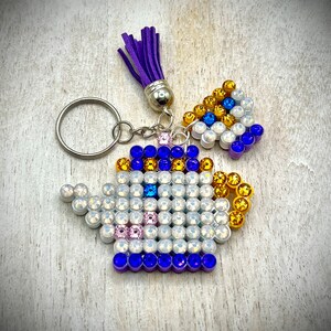 May include: A decorative keychain featuring a teapot design, embellished with iridescent and colorful rhinestones. The teapot is accented with blue, gold, and pink stones, and has a purple tassel. The keychain includes a silver keyring.