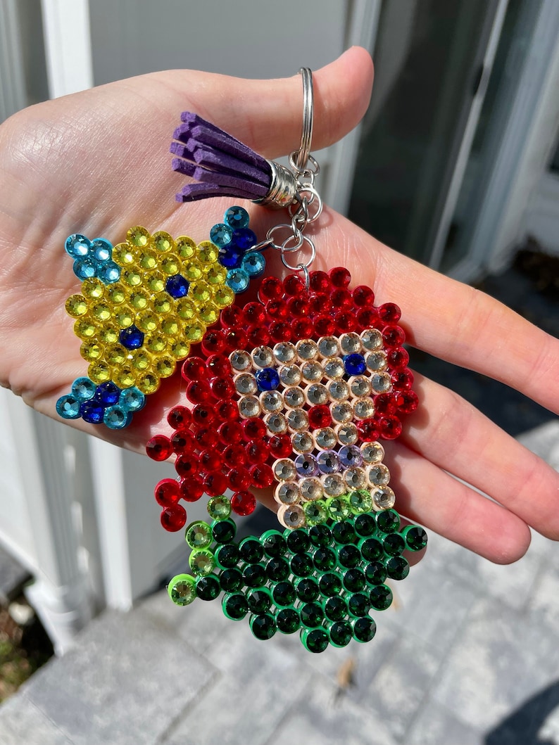 Puede incluir: Un llavero hecho a mano con una sirena pelirroja y un pez amarillo, ambos adornados con pedrer&iacute;a de colores. La sirena tiene una cola verde. El llavero incluye un anillo plateado y una borla morada.