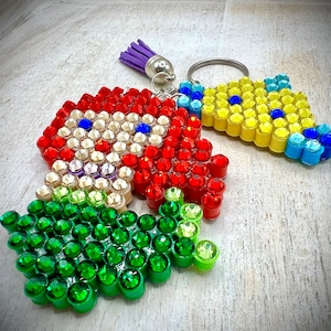 Puede incluir: Tres llaveros con adornos coloridos en forma de joyas. Uno es rojo con detalles blancos y azules, otro es amarillo y azul, y el tercero es verde. Cada llavero tiene un anillo plateado y una borla morada.