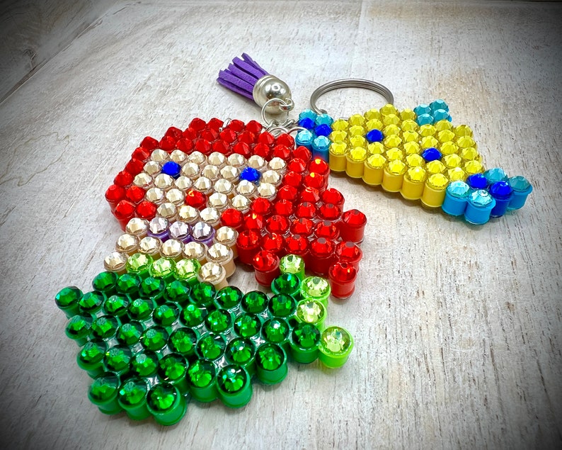 Puede incluir: Tres llaveros con adornos coloridos en forma de joyas. Uno es rojo con detalles blancos y azules, otro es amarillo y azul, y el tercero es verde. Cada llavero tiene un anillo plateado y una borla morada.
