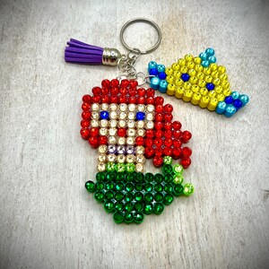 Puede incluir: Un llavero con una sirena pelirroja y un pez amarillo, ambos adornados con brillantes pedrer&iacute;a. La sirena tiene una cola verde y el pez tiene detalles azules. Una borla morada cuelga del anillo del llavero.