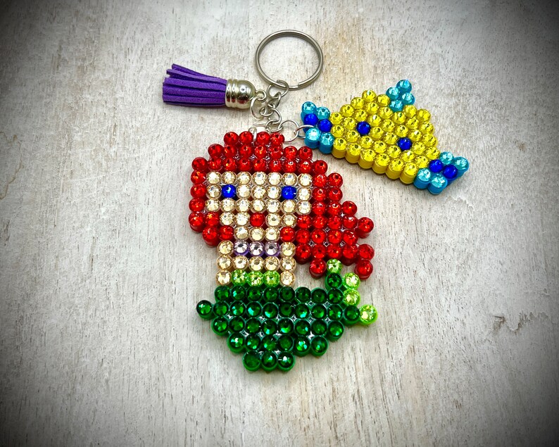 Puede incluir: Un llavero con una sirena pelirroja y un pez amarillo, ambos adornados con brillantes pedrer&iacute;a. La sirena tiene una cola verde y el pez tiene detalles azules. Una borla morada cuelga del anillo del llavero.