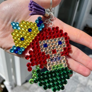 Puede incluir: Un llavero con un dise&ntilde;o de cuentas de un personaje de dibujos animados con pelo rojo y un pez amarillo. El personaje tiene el pelo rojo y el pez es amarillo. El llavero tiene un anillo plateado y una borla morada.