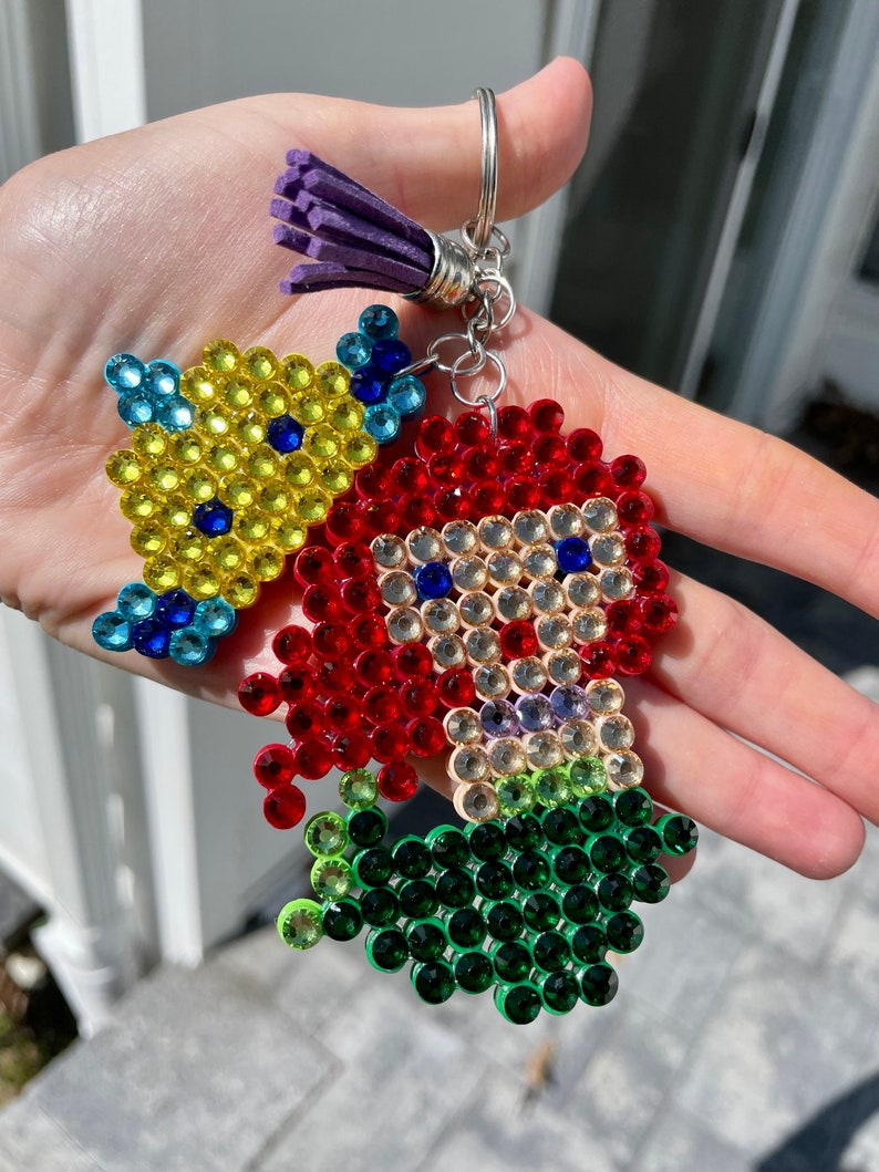 Puede incluir: Un llavero con un dise&ntilde;o de cuentas de un personaje de dibujos animados con pelo rojo y un pez amarillo. El personaje tiene el pelo rojo y el pez es amarillo. El llavero tiene un anillo plateado y una borla morada.