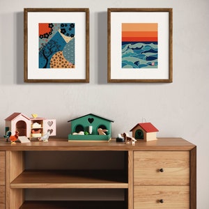 Puede incluir: Dos impresiones enmarcadas en una pared blanca, una con un dise&ntilde;o geom&eacute;trico azul y naranja y la otra con un dise&ntilde;o de olas azul y naranja. Debajo de las impresiones hay una estanter&iacute;a de madera con dos cajones y dos estantes. Hay tres casas de juguete de madera en la estanter&iacute;a.