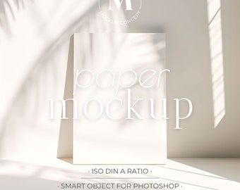 Peut inclure: Une maquette de papier blanche avec le texte "paper mockup" dessus. Le papier est sur une surface blanche avec des ombres d'une fenêtre et d'une plante. Le texte "ISO DIN A RATIO", "SMART OBJECT FOR PHOTOSHOP", et "PSD & JPG FILES" est en dessous du papier.