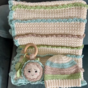 May include: A handmade crochet baby set in cream, green, blue, and brown. The set includes a blanket, a hat, and a toy with a wooden ring. The blanket and toy have scalloped edges. The hat has horizontal stripes.