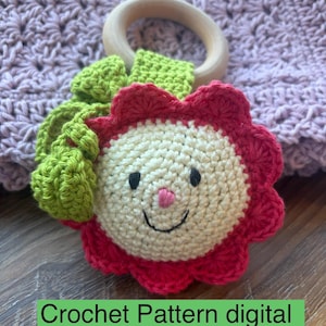 May include: A handmade crochet baby toy in the shape of a smiling flower with a wooden ring. The flower has a cream-colored center, red petals, and green leaves. The text "Crochet Pattern digital download" is on the image.